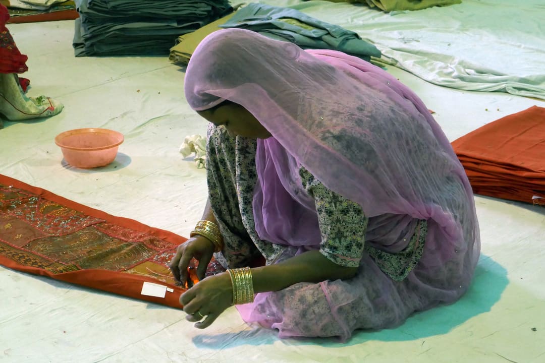 A woman in a pink dupatta sits on the floor of a workshop stitching an embroidered red textile runner.