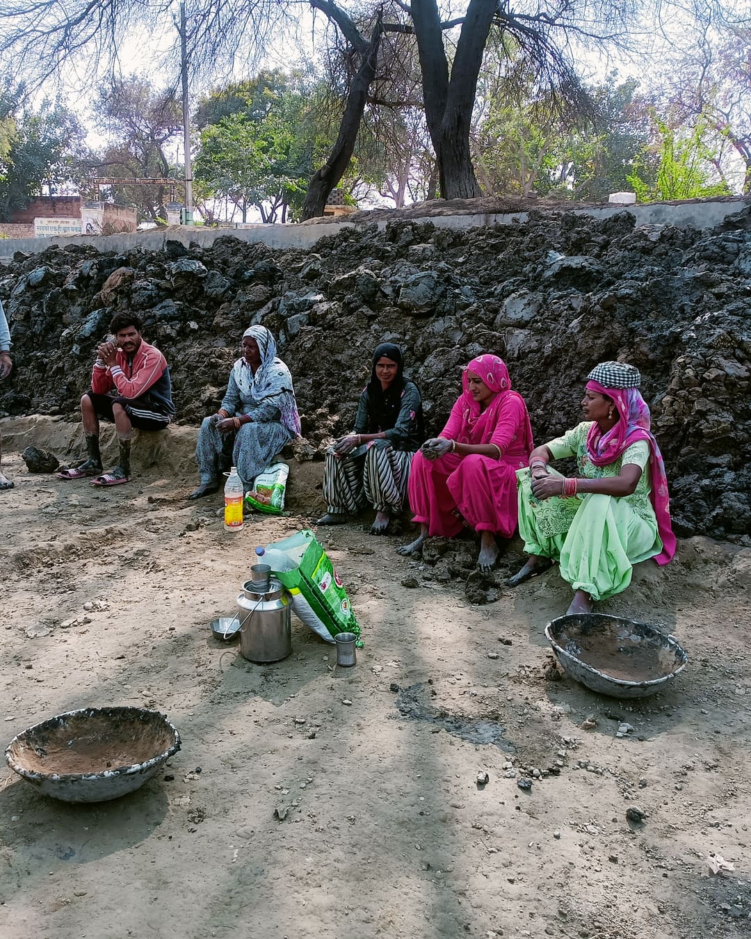 A group of women in bright salwar suits and dupattas sit on the ground beside a stone wall with iron pans, resting during public works.