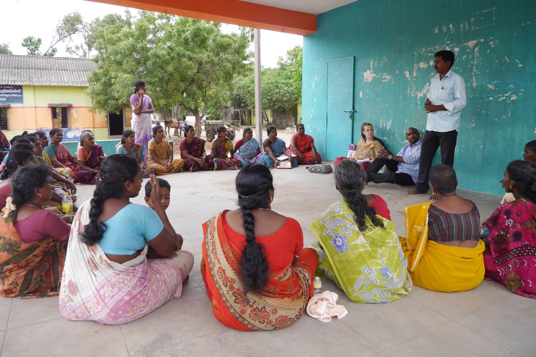 Women sit in a circle on the floor of a covered community hall while a man stands to address them at a self-help group meeting.